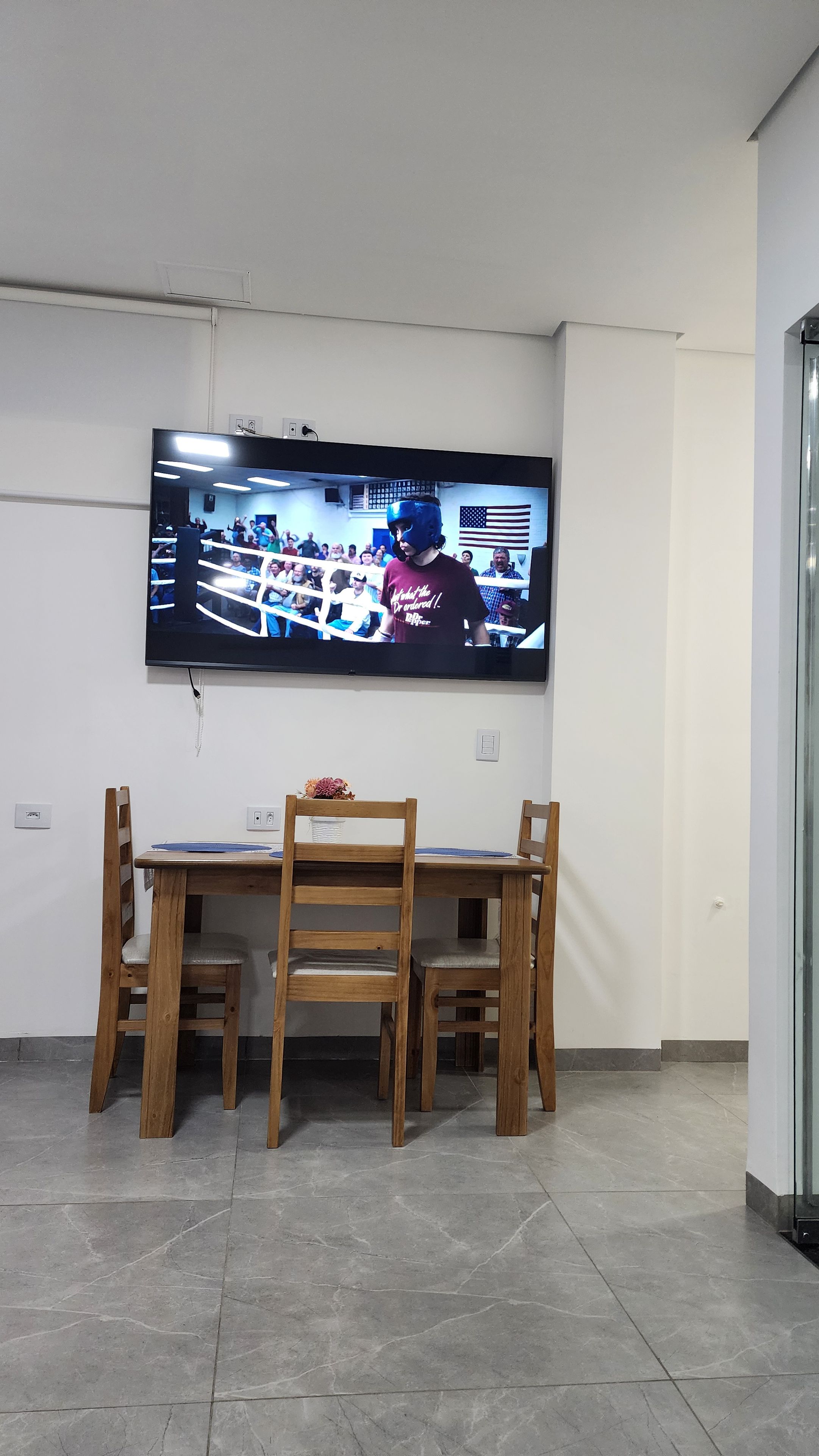 Een kamer met een wandgemonteerde flatscreen televisie, een houten eettafel met stoelen en een betegelde vloer.