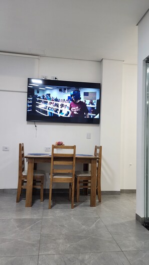 A room with a flat-screen TV mounted on the wall, a wooden dining table with chairs, and a tiled floor.