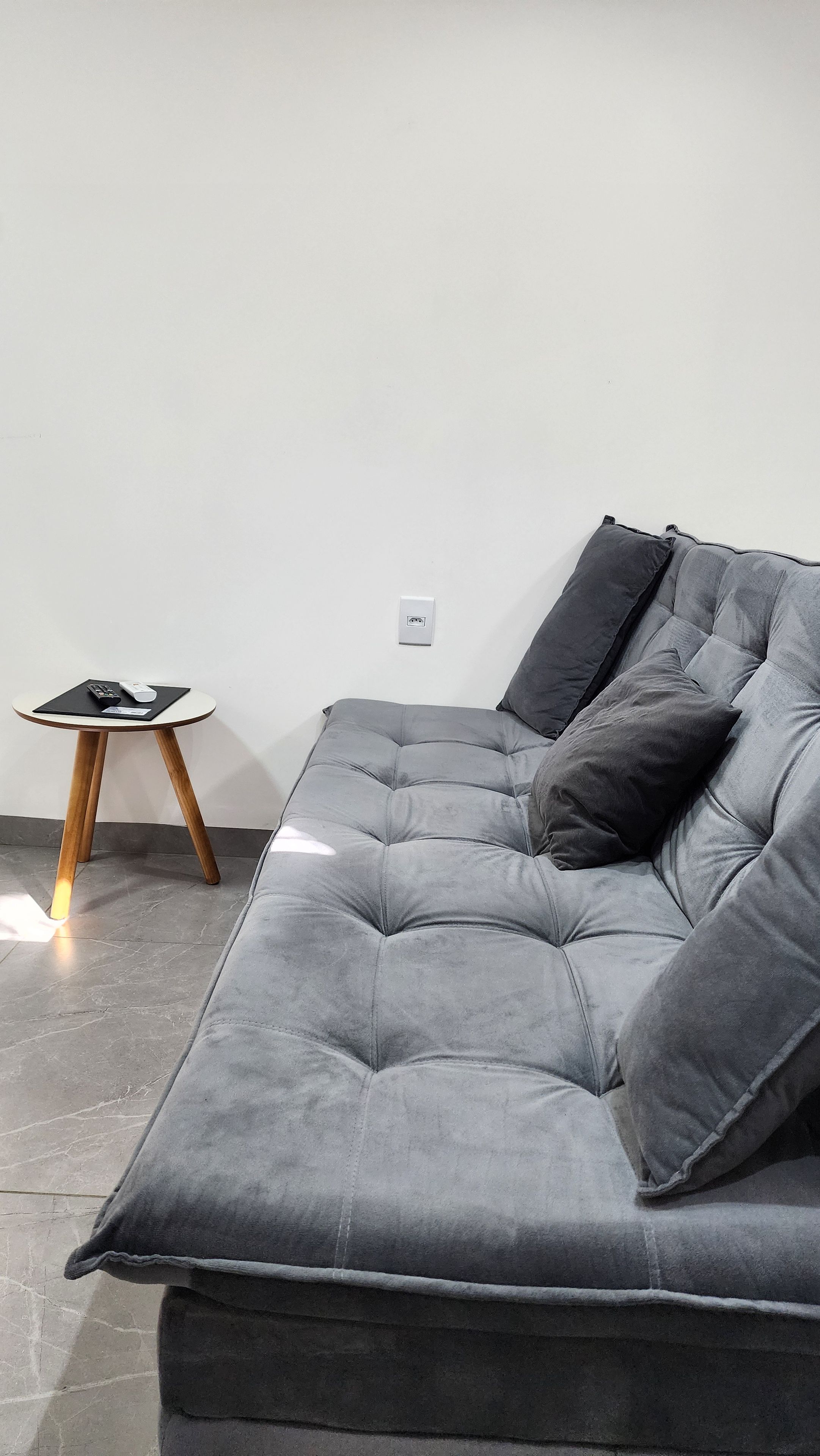 Un sofá gris con un cojín, una mesita de madera con un control remoto y un enchufe en la pared.
