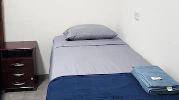 Ein Einzelbett mit grauem Matratze und blauer Decke, ein Nachttisch aus Holz mit Schubladen sowie ein kleiner Schreibtisch mit Stuhl.