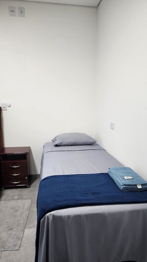 A single bed with a grey mattress and a blue blanket, a wooden nightstand with drawers, and a small desk with a chair.