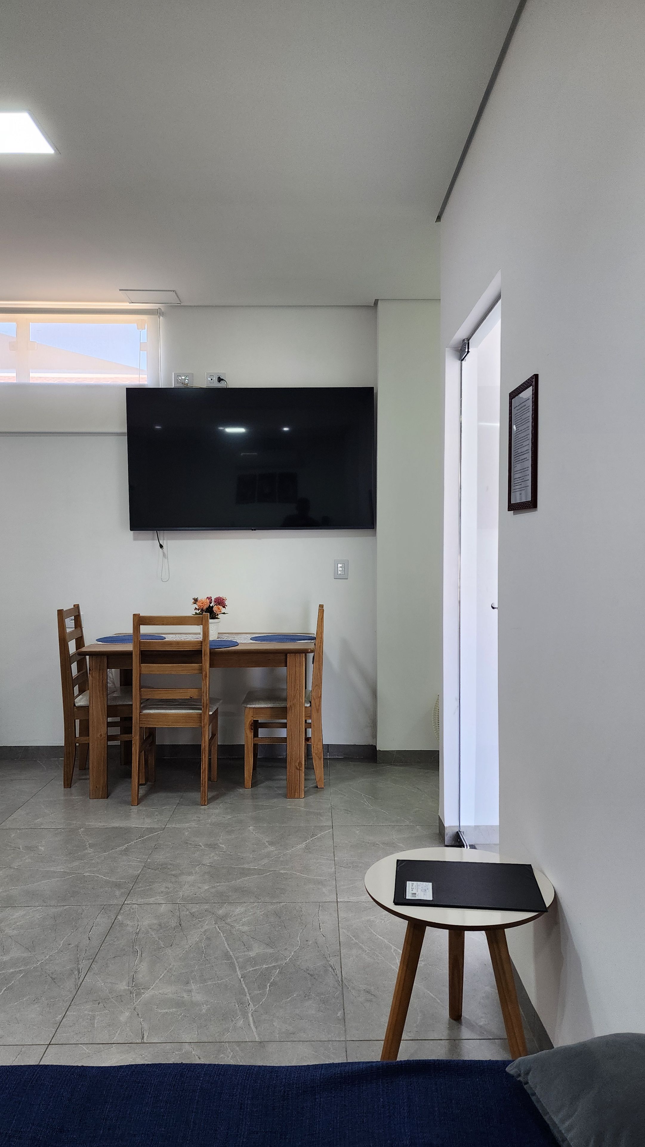A room with a flat-screen TV mounted on the wall, a dining table with chairs, a small round table with a laptop, and a door with a framed item on the wall.
