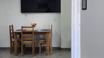 A room with a flat-screen TV mounted on the wall, a dining table with chairs, a small round table with a laptop, and a door with a framed item on the wall.