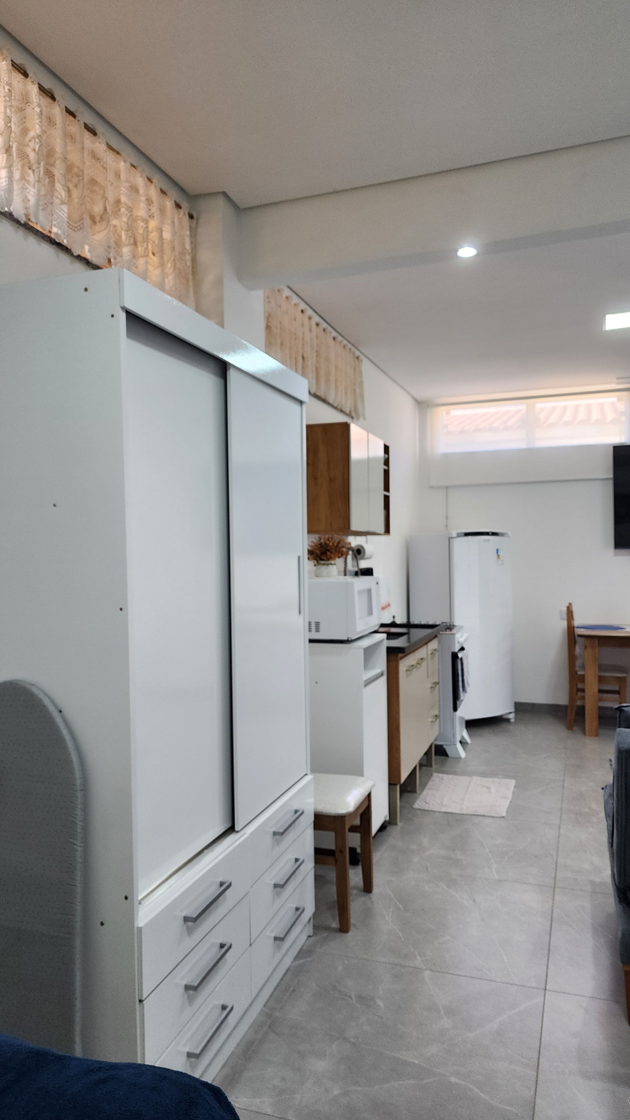 A compact kitchen with white appliances, a wooden cabinet, and a dining area with a chair.