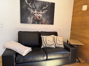 A black leather sofa with a white throw blanket and patterned pillows, a small wooden side table, and a painting of a deer on the wall.