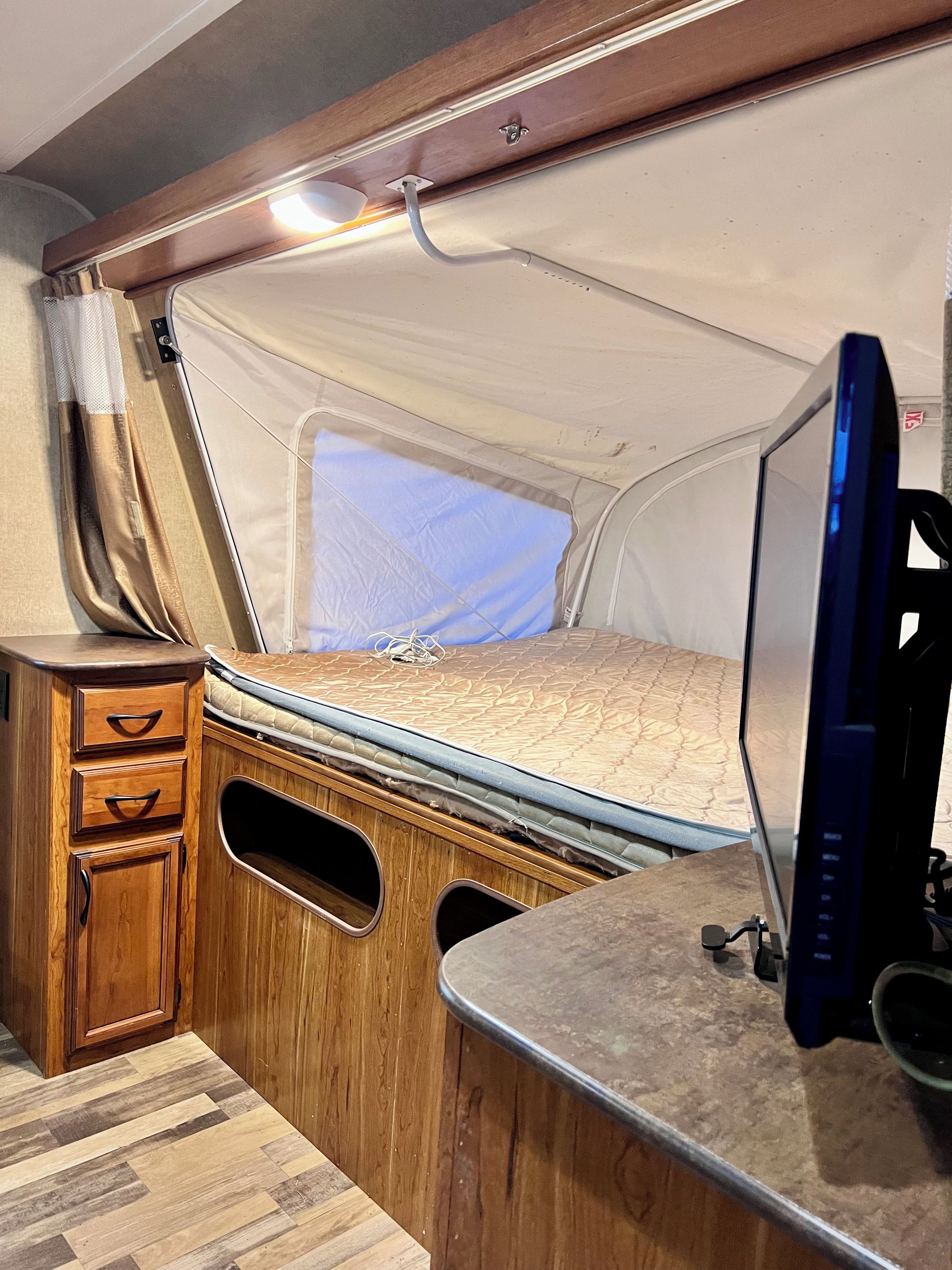 A compact RV interior with a bed, a wooden cabinet with drawers, a television, and a window.