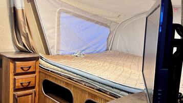 A compact RV interior with a bed, a wooden cabinet with drawers, a television, and a window.