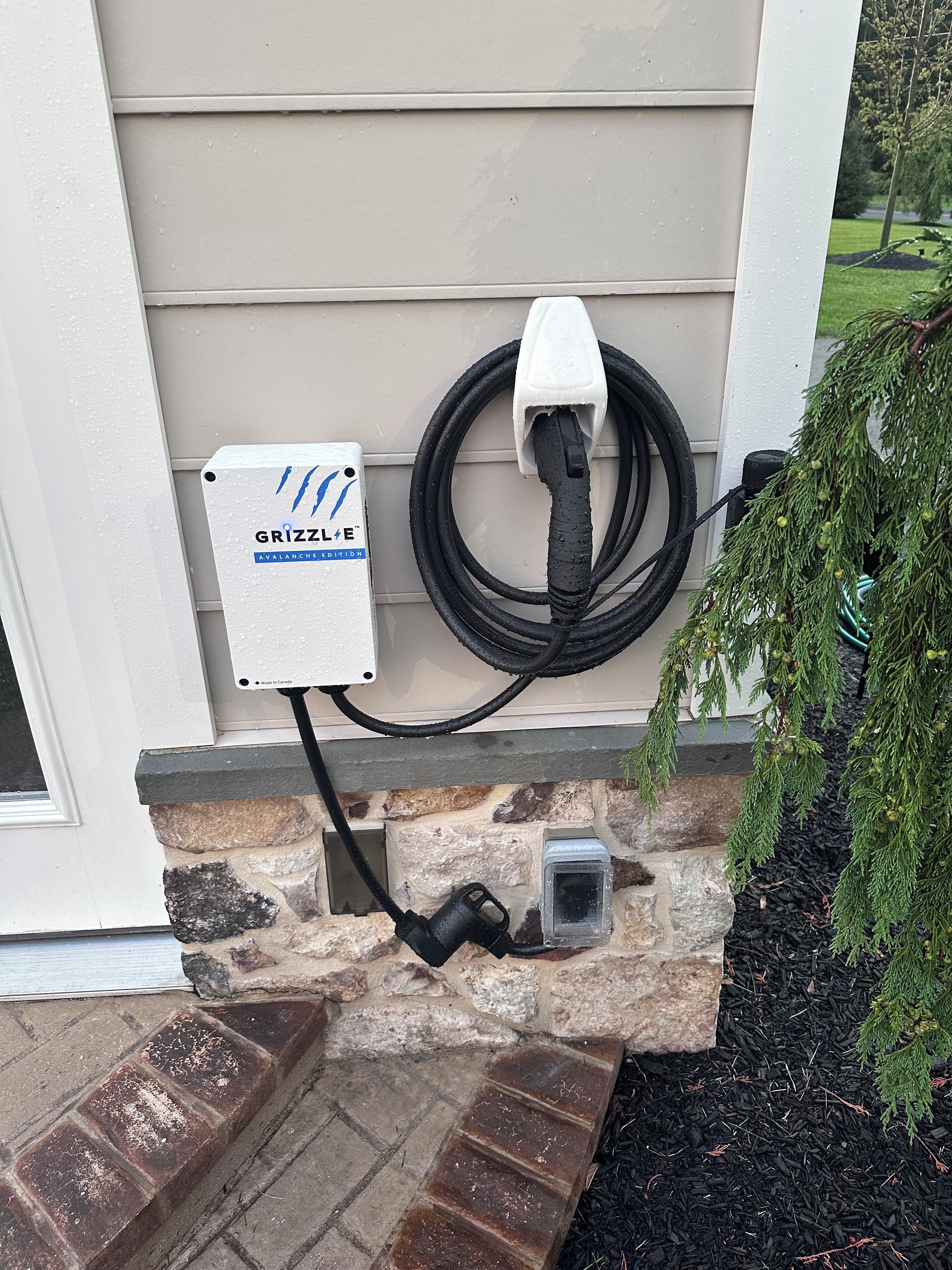 An outdoor electrical outlet with a cord plugged in, a white box labeled 'GRIZZLE AVALANCHE EDITION,' and a stone wall.