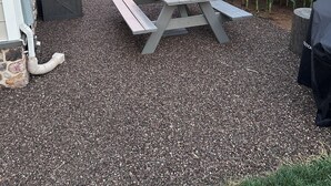Uma área de pátio com mesa de piquenique, cadeiras e guarda-sol, cercada por chão de brita e um milharal.