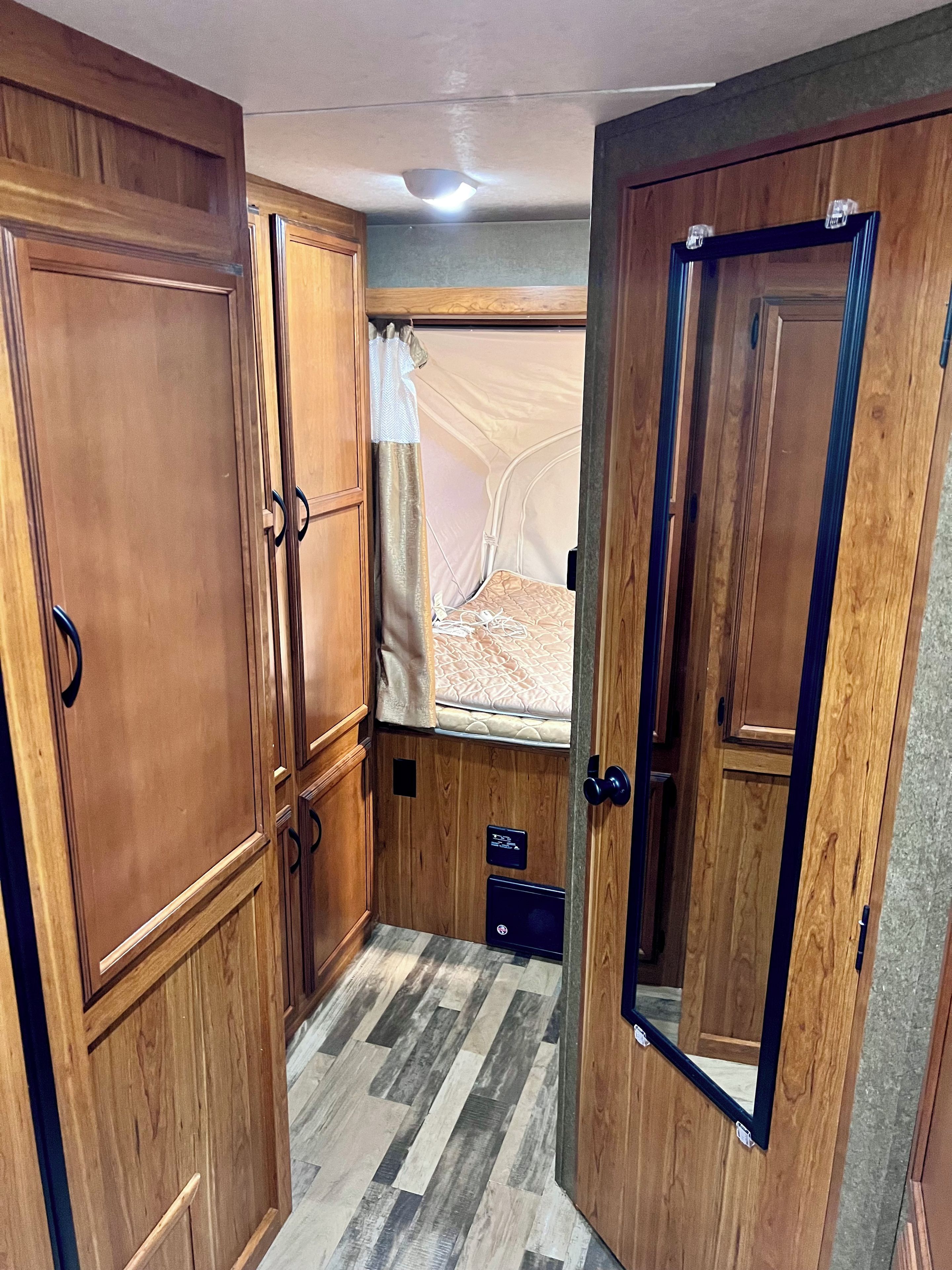 A compact room with wooden cabinetry, a bed with a mattress and pillows, a window with curtains, and a small storage unit.