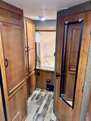 Quarto compacto com armários de madeira, cama com colchão e almofadas, janela com cortinas e um pequeno móvel para guardar objetos.