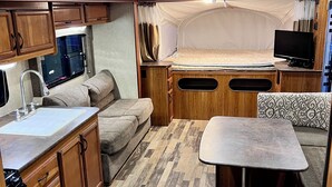 Interior compacto de um veículo recreativo, com cozinha compacta, cama, área de estar e televisão.