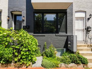 Ein modernes, zweistöckiges Haus mit einer dunkelgrauen Fassade, einem großen Fenster und einem kleinen Garten mit verschiedenen Pflanzen.