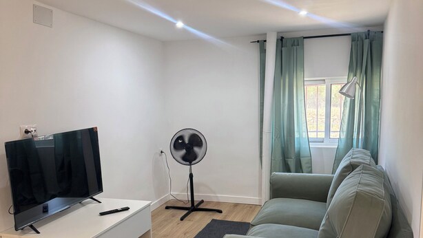 A modern living room with a flat-screen TV, a standing fan, a sofa, and a window with curtains. - Cozy 1-bedroom apartment in lovely Ambite with beutiful views (Ambite)