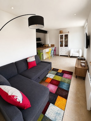 A modern living room with a grey sofa, colorful rug, and a kitchen area in the background.