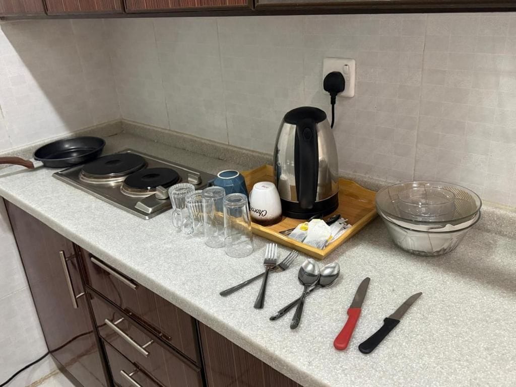 A kitchen counter with a kettle, a frying pan, a cutting board, and various utensils.