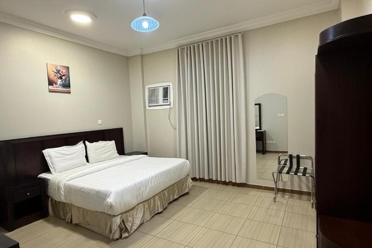 Habitación de hotel con cama, mesita de noche, espejo, ventana con cortinas y aire acondicionado.