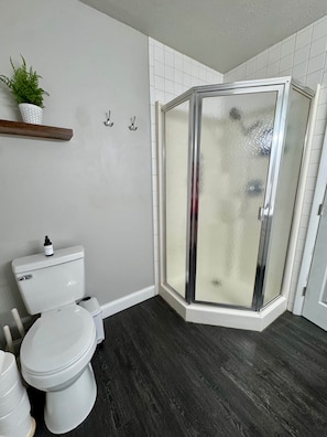 Un baño con inodoro, ducha con puerta de cristal, lavamanos blanco y una planta en un estante.