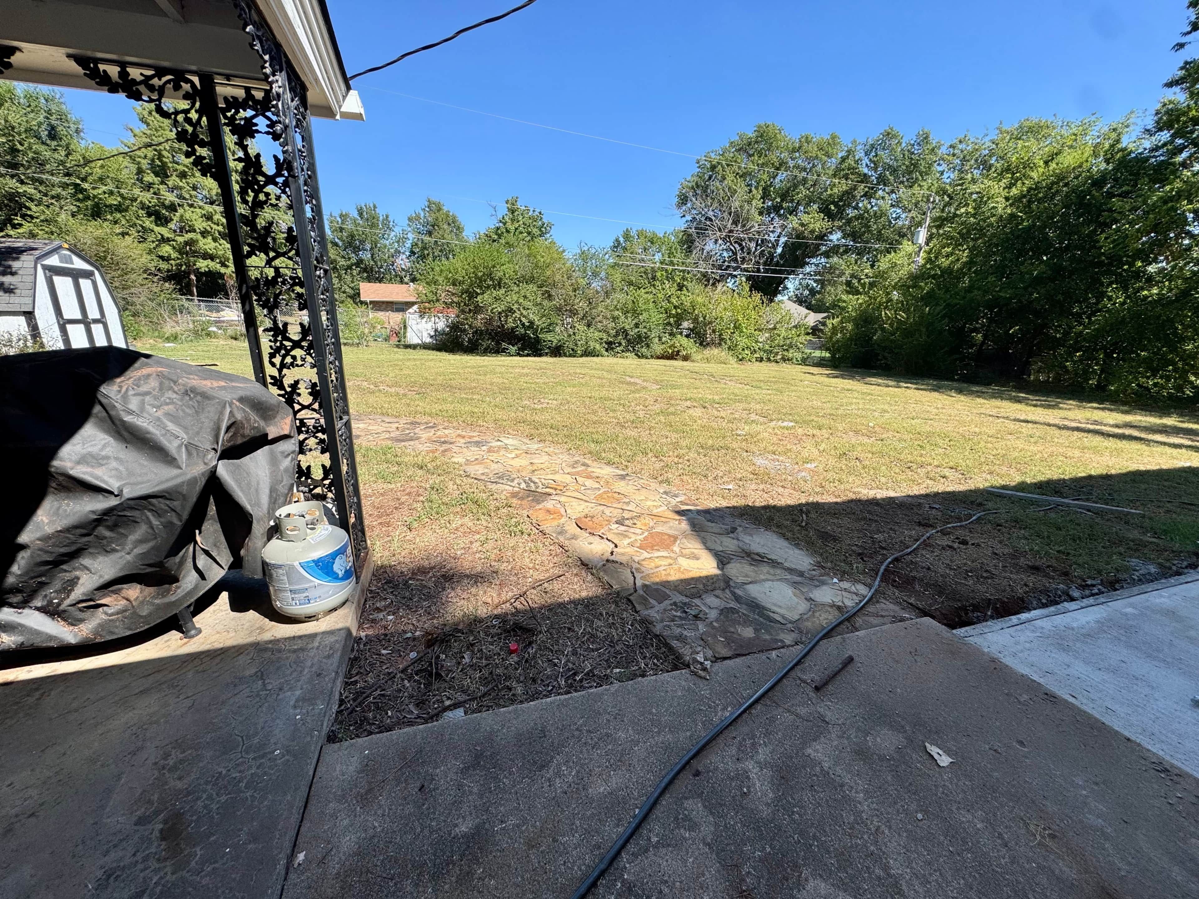 A backyard with a stone pathway leading to a house, a covered grill, and a hose on the ground.
