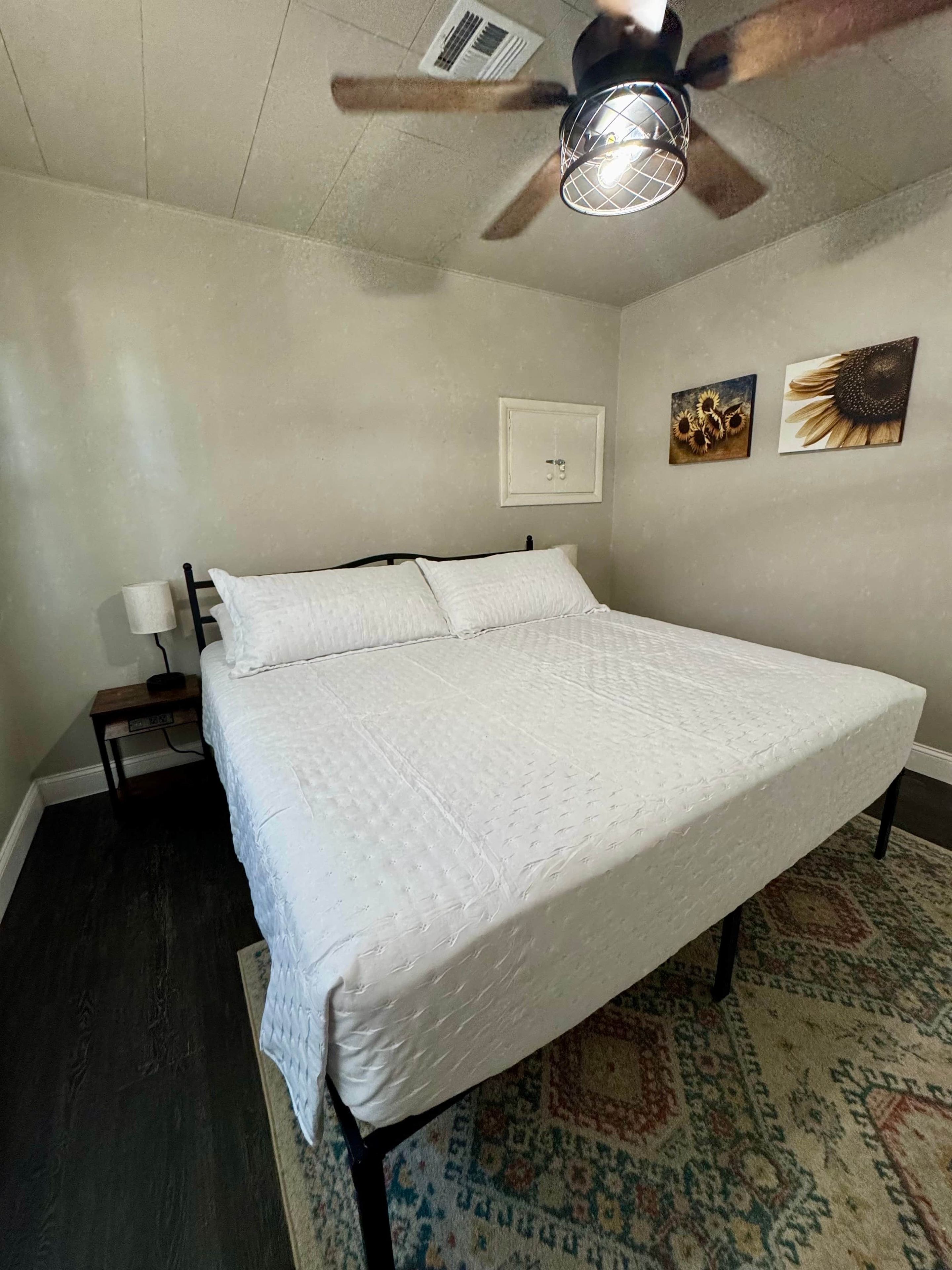 Un dormitorio con cama, ventilador de techo, mesita de noche, interruptor de luz en la pared y dos cuadros decorativos.