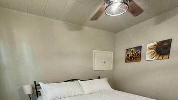 Un dormitorio con cama, ventilador de techo, mesita de noche, interruptor de luz en la pared y dos cuadros decorativos.