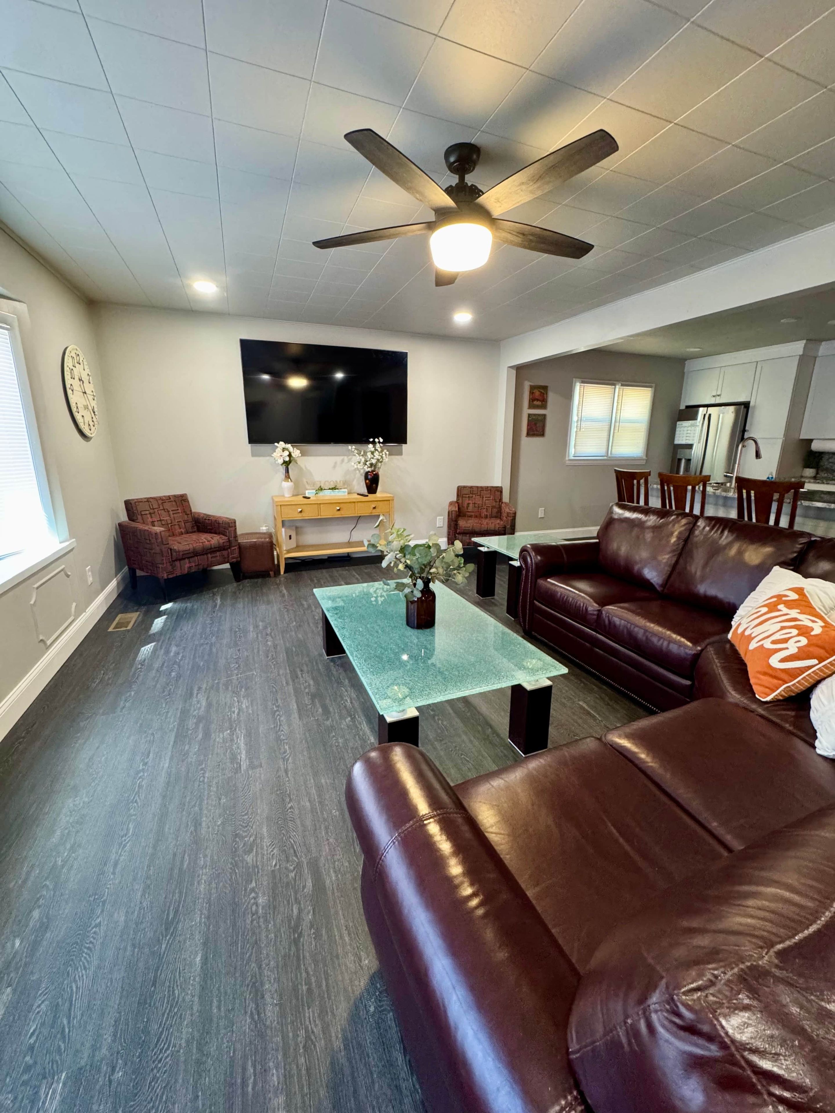 A living room with a brown leather couch, a glass coffee table, a television mounted on the wall, and a ceiling fan.