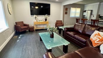 A living room with a brown leather couch, a glass coffee table, a television mounted on the wall, and a ceiling fan.