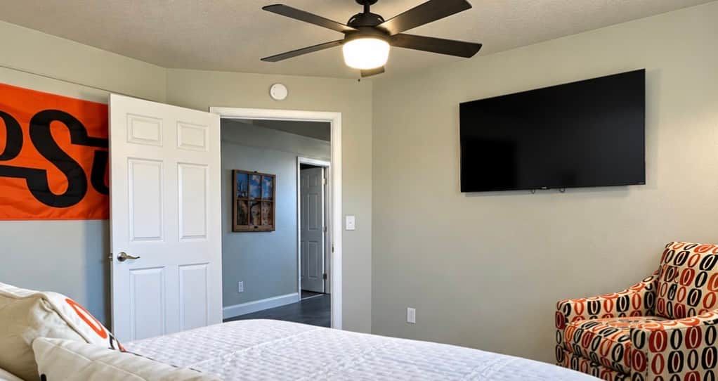 A bedroom with a bed, a ceiling fan, a TV mounted on the wall, and a patterned armchair.