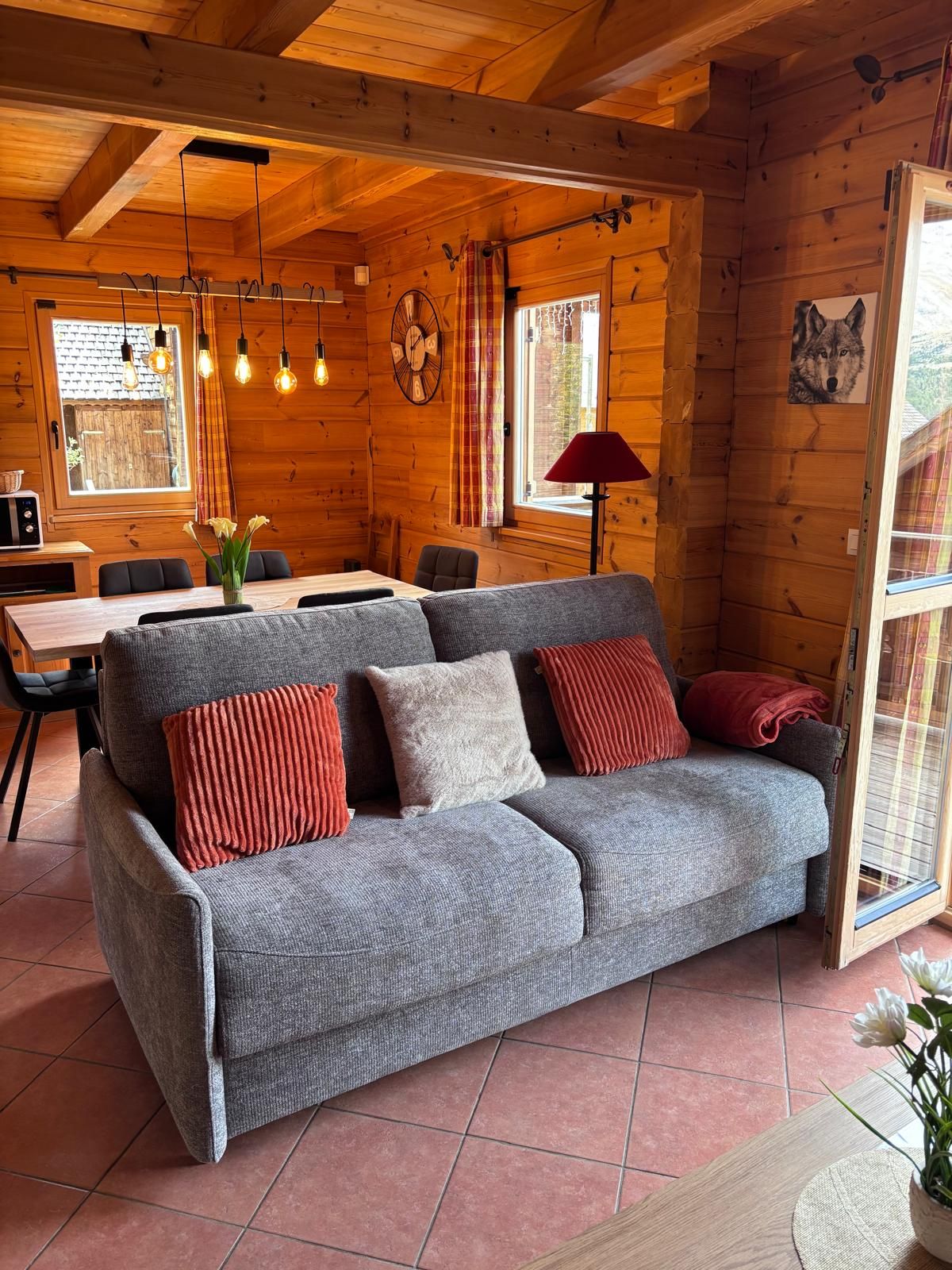 A cozy cabin interior with a grey sofa, red and white cushions, a dining table, and a large window with curtains.