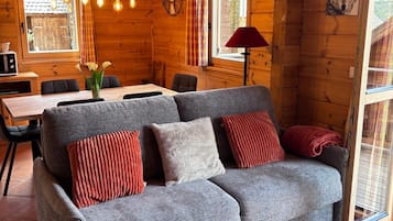 L’intérieur d’un chalet accueillant, avec un canapé gris, des coussins rouges et blancs, une table à manger et une grande fenêtre agrémentée de rideaux.