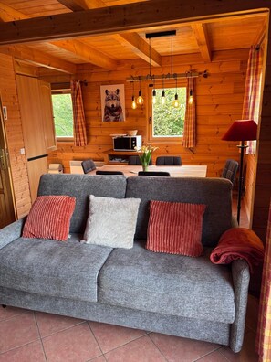 A cozy cabin interior with a grey sofa, wooden walls, a dining area, and a window with curtains.
