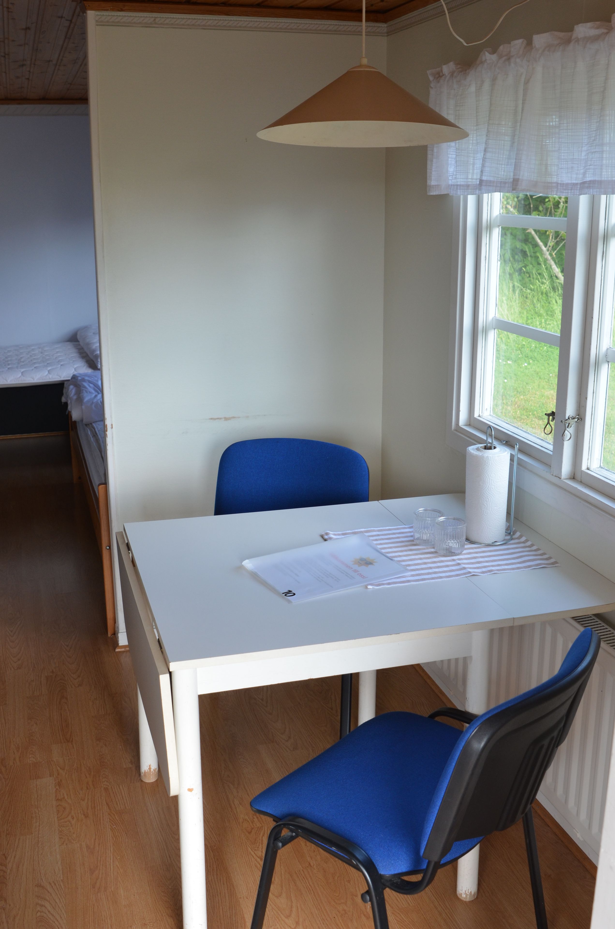A room with a white desk, a blue chair, a bed, and a window with a view of greenery.