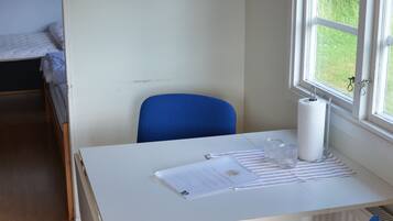 A room with a white desk, a blue chair, a bed, and a window with a view of greenery.