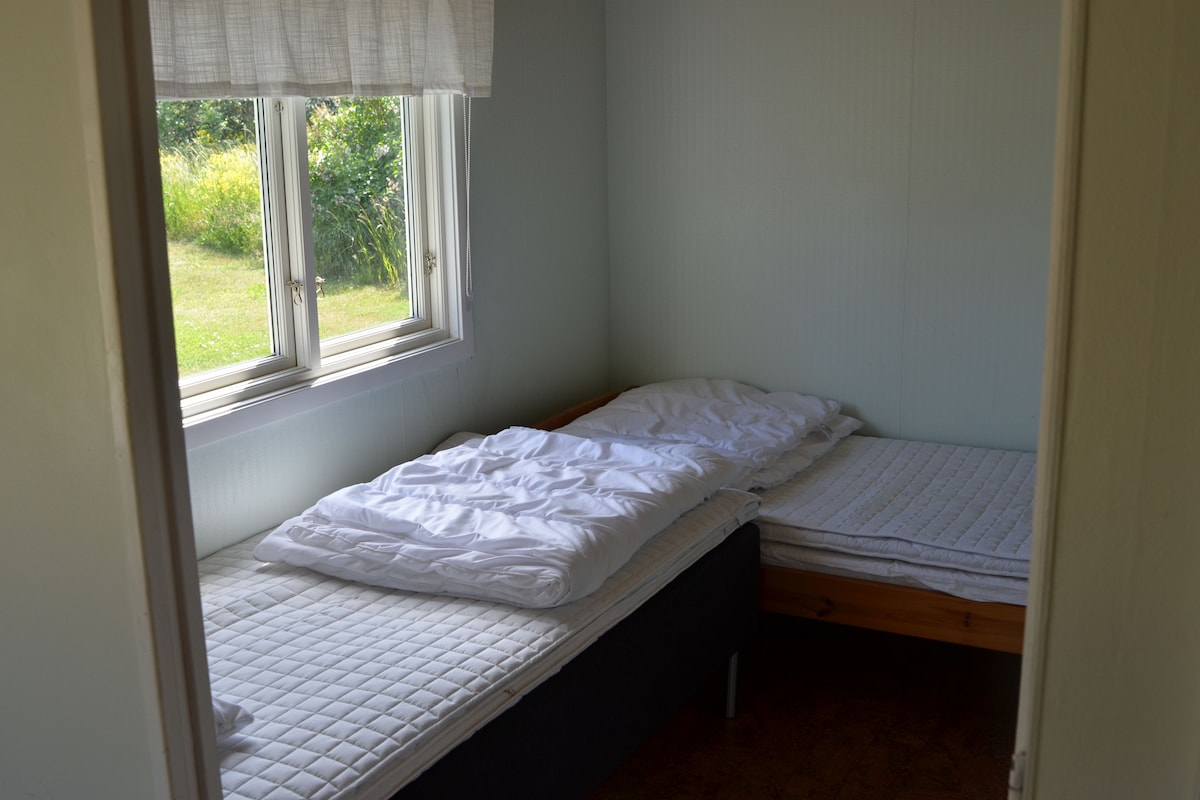 A small, single-bed room with a window, a door, and a view of greenery outside.