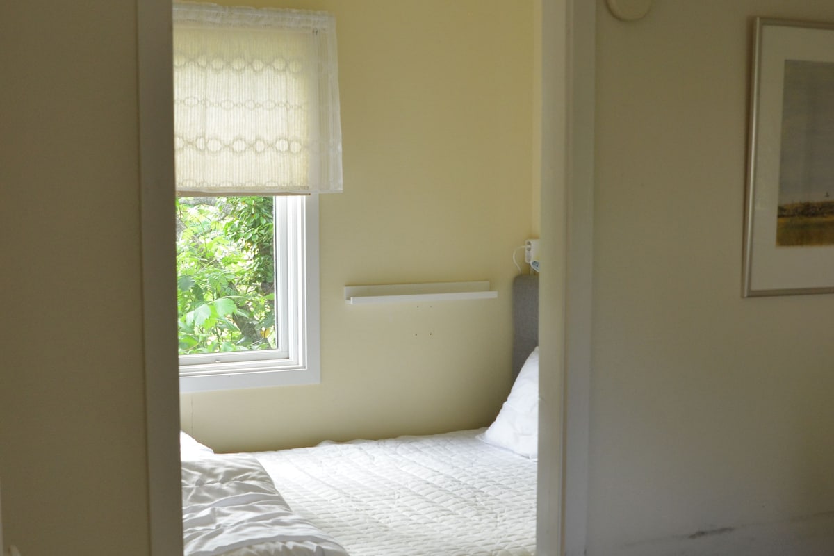 A small, single-bed room with a window, a bedside table, and a wall-mounted light fixture.
