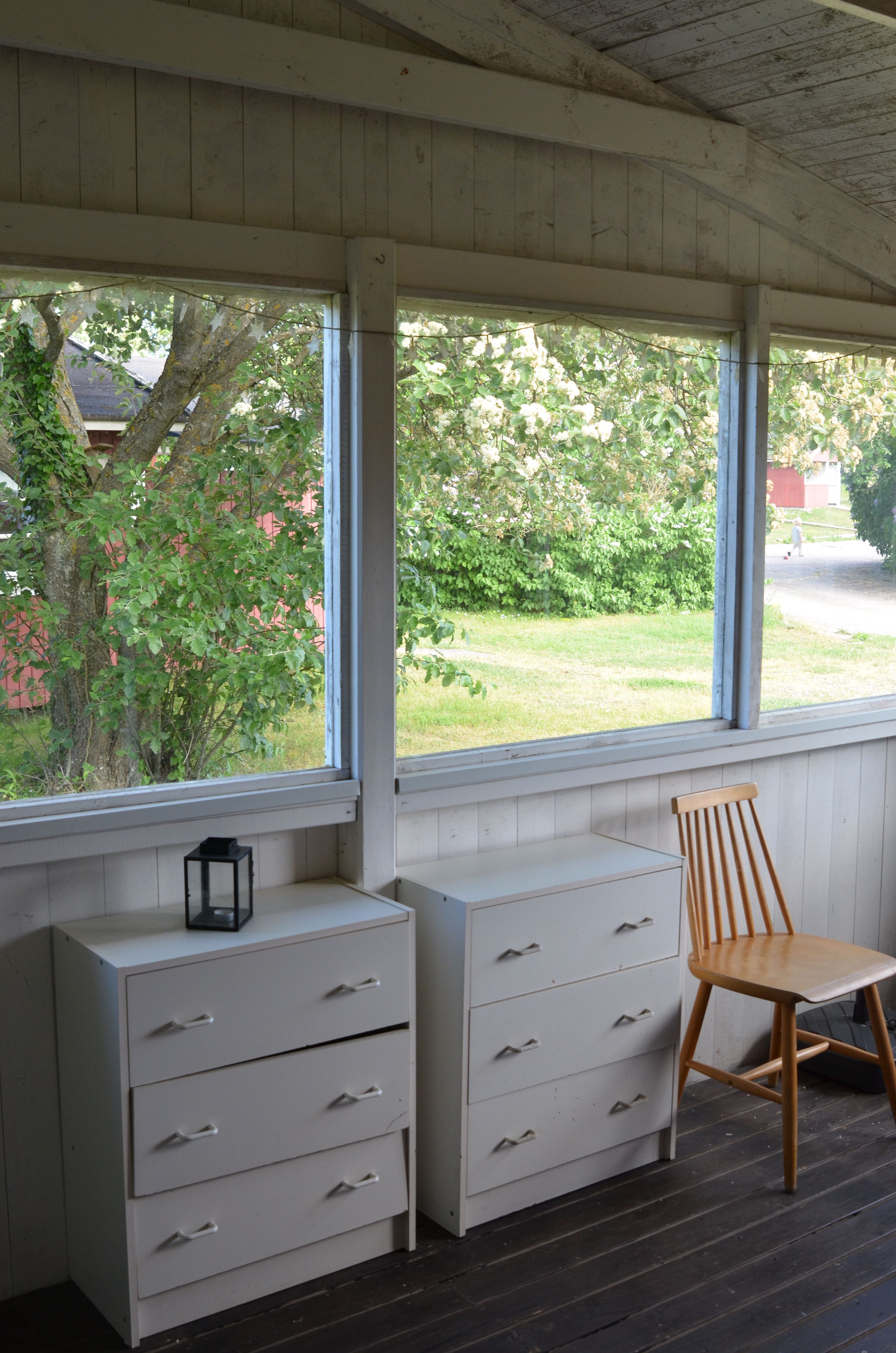 A room with a wooden ceiling, a window with a view of greenery, a wooden chair, and a white dresser with drawers.