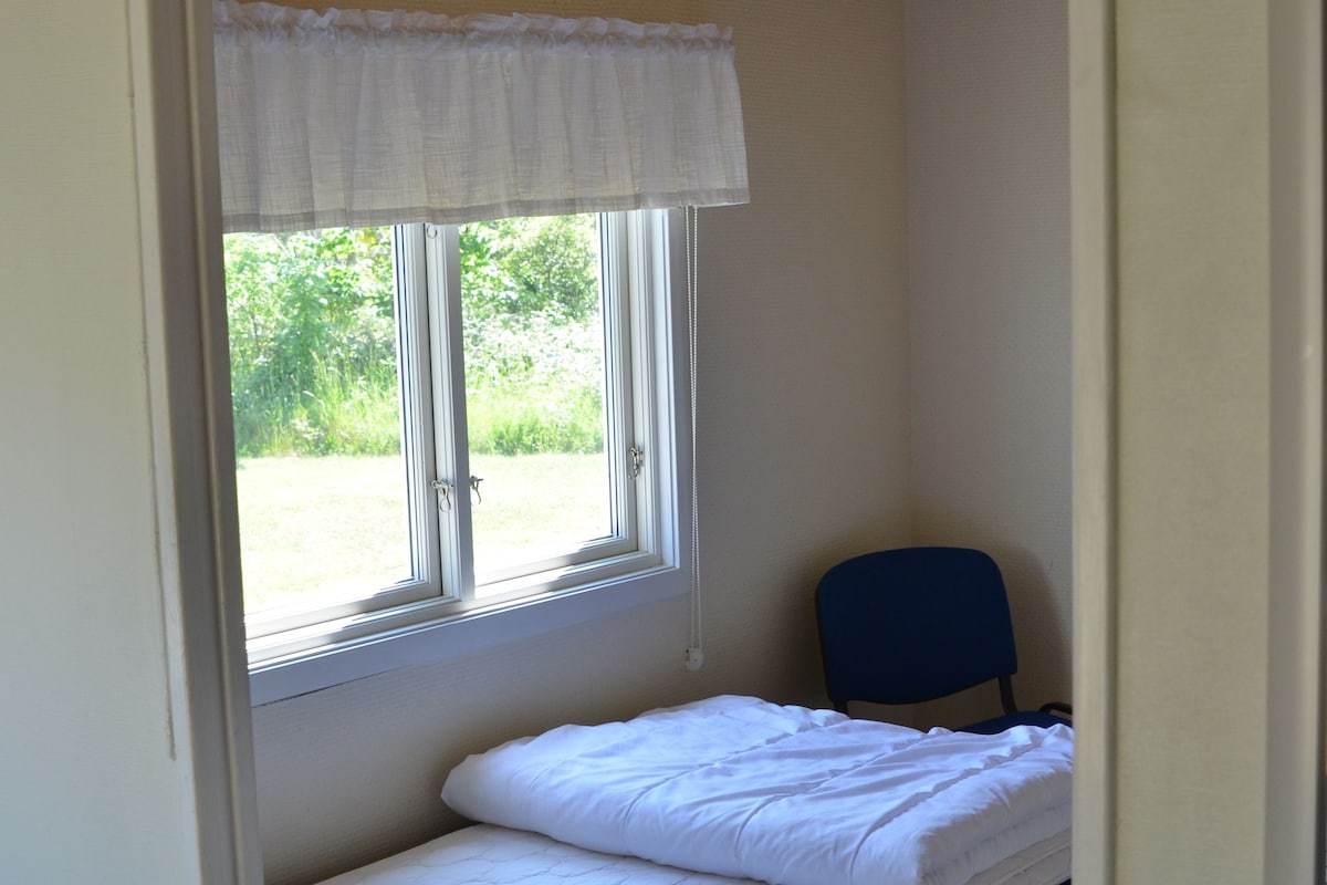 A small, single-bed room with a window, a chair, and a wooden ceiling.