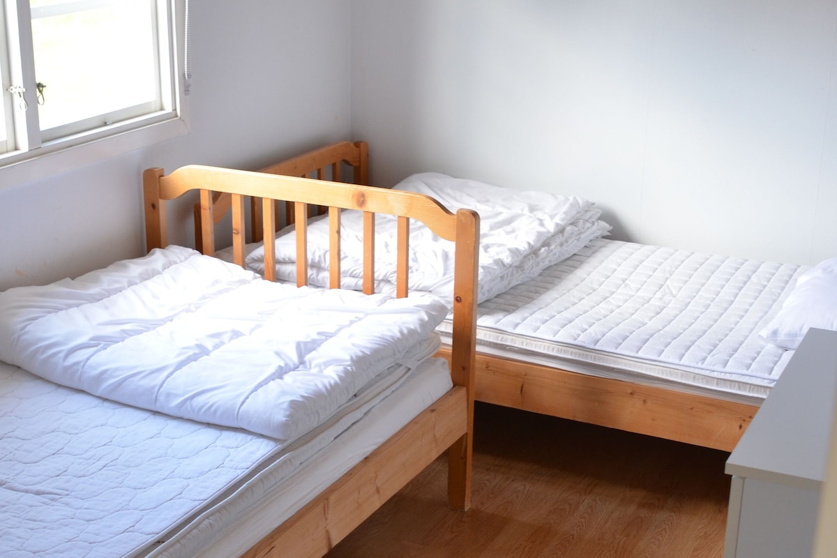 A small, single-room accommodation with two wooden bunk beds, a window with a sheer curtain, and a white wardrobe.