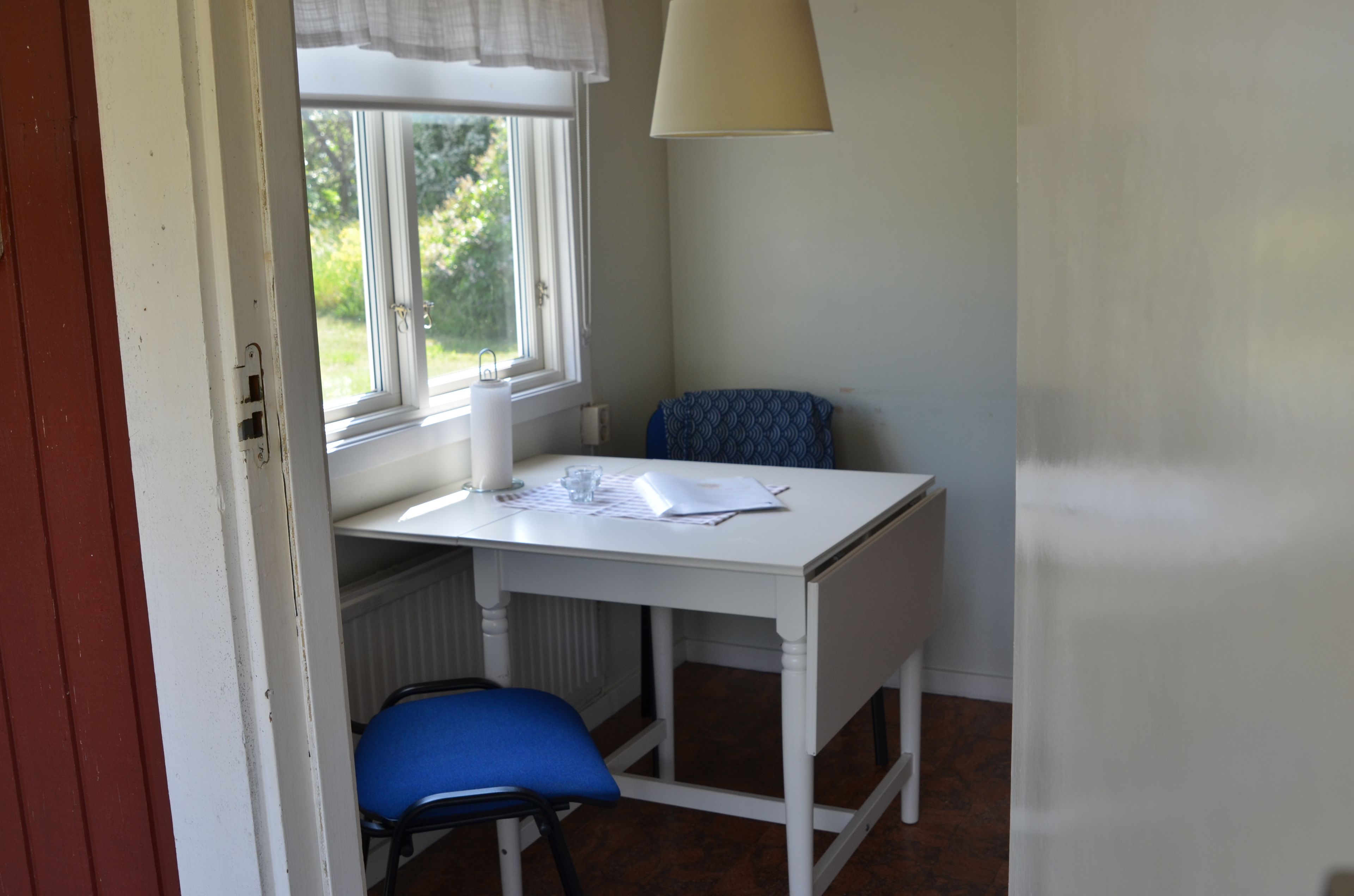 Un escritorio pequeño y blanco con una silla, un ventanal con vistas a la vegetación y una lámpara.
