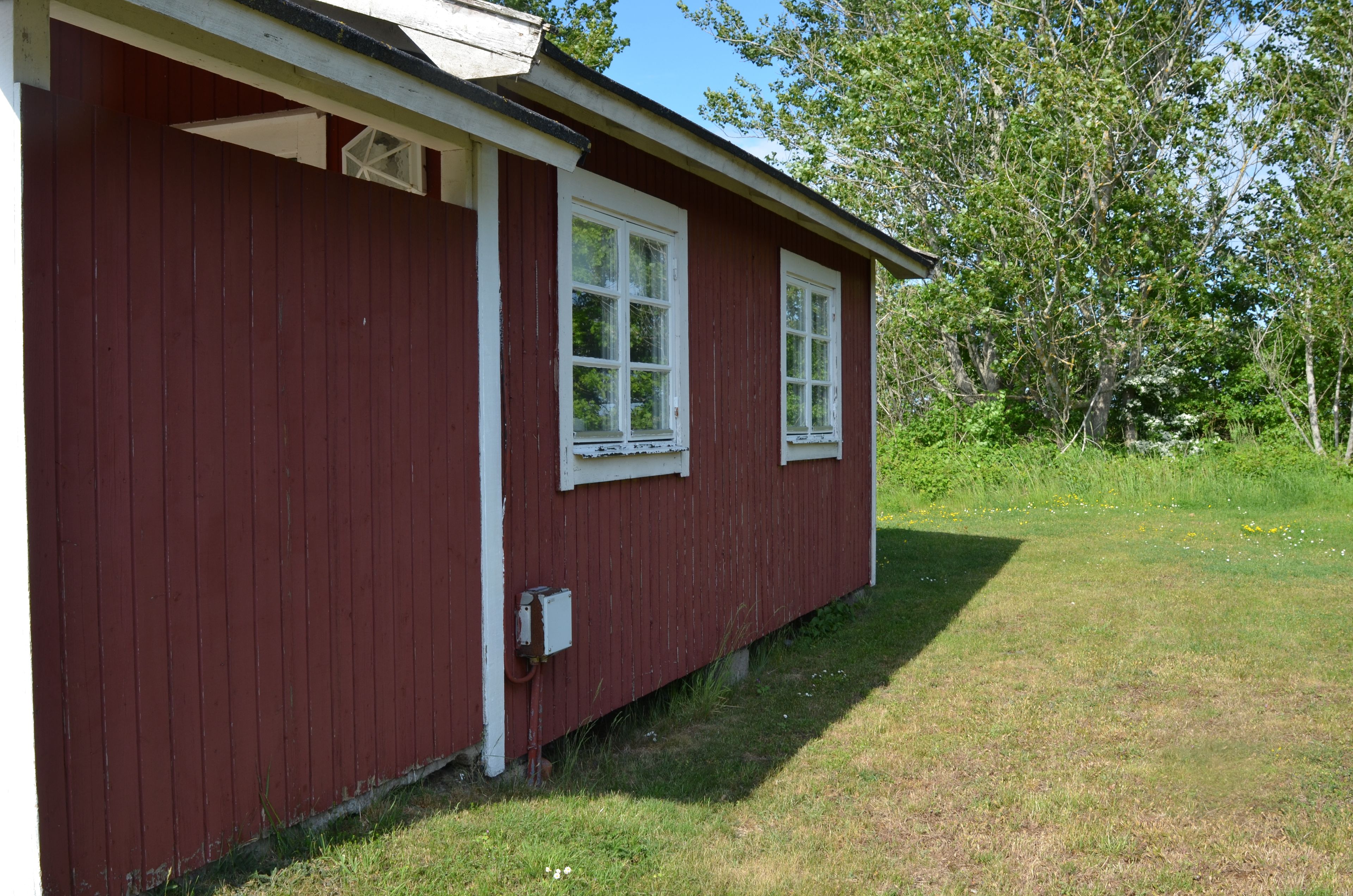 A red wooden building with two windows and a small electrical box on the side.