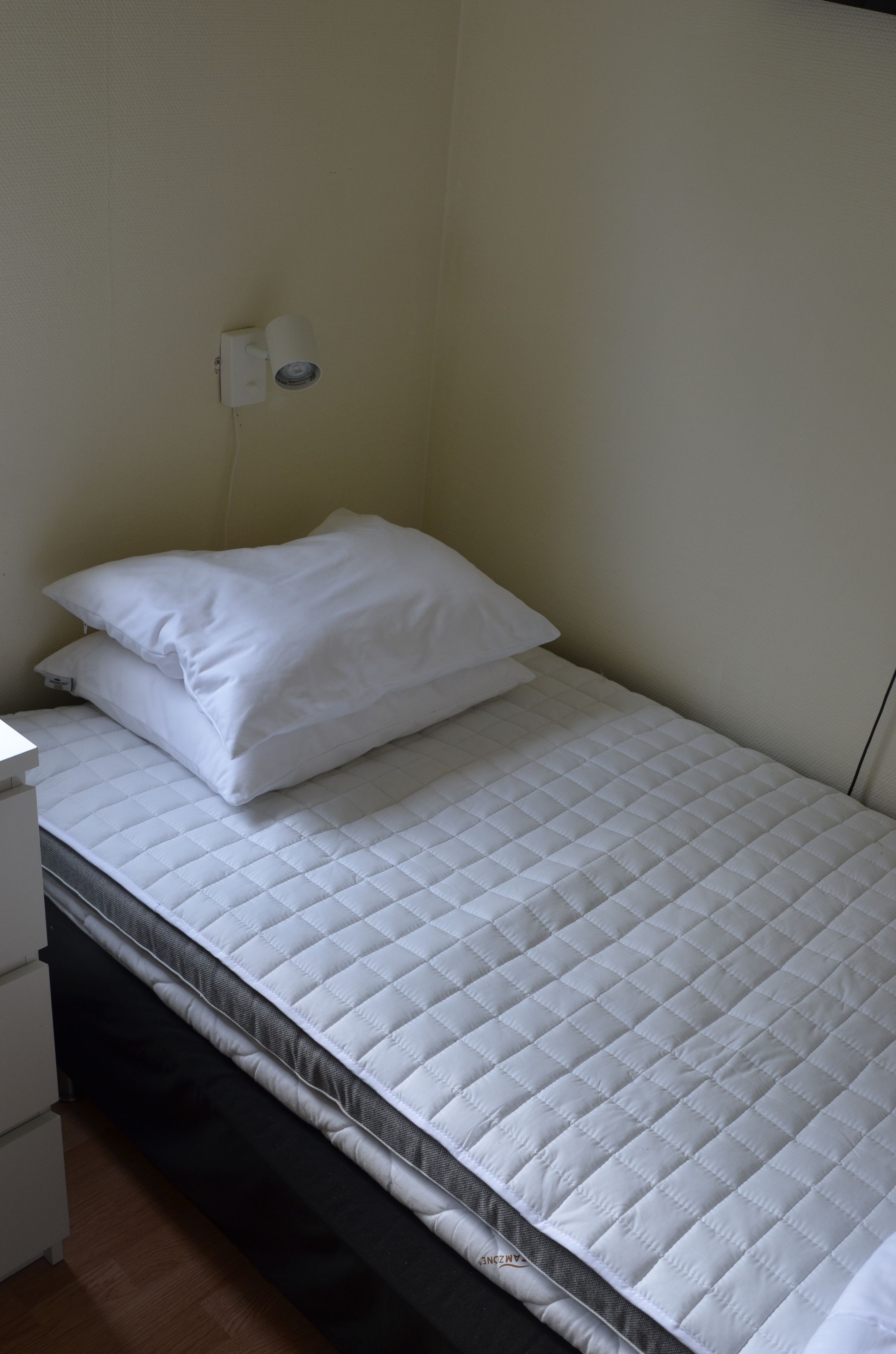 Una cama individual con colchón y almohadas blancos, una mesita de noche pequeña y una lámpara de pared.