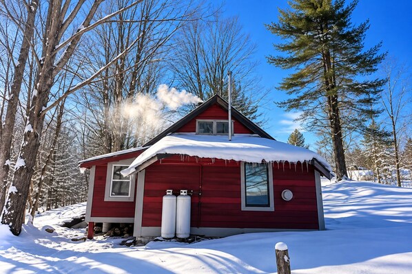 Une petite cabane rouge, dont le toit est recouvert de neige, entourée d’arbres et de neige.