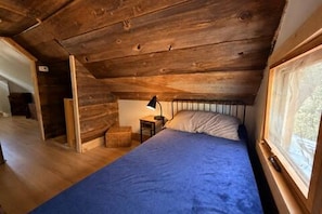 Un lit simple recouvert d’une couverture bleue, dans une pièce dont le plafond est orné de poutres en bois et qui possède une fenêtre.