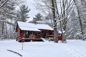 Une cabane dont le toit est recouvert de neige, entourée d’arbres, avec une pile de bois de chauffage devant.