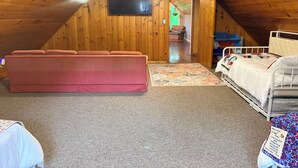 Une chambre confortable située sous les toits, avec un plafond en pente en bois, une grande fenêtre, un téléviseur fixé au mur, un canapé rose, un lit blanc et un tapis coloré.