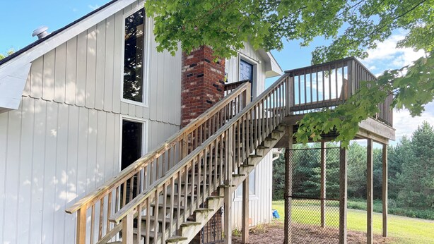 Un escalier en bois mène à un espace extérieur abrité, doté d’une cheminée en briques et d’une porte bleue.