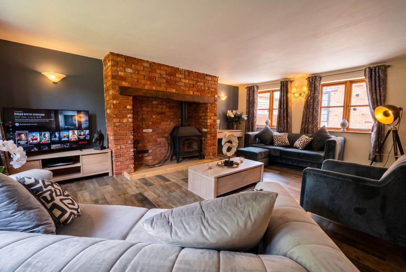 A cozy living room with a brick fireplace, a flat-screen TV, comfortable seating, and a large window with curtains.