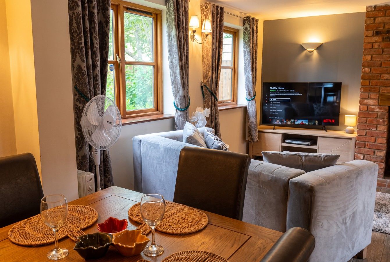 Un salon confortable avec une télévision, une table à manger dressée avec des sets de table et des verres, et un ventilateur près d’une fenêtre.