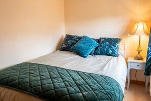 A bed with a quilted green bedspread and blue pillows, a bedside table with a lamp, and a wall with a warm light source.