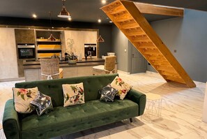 Een moderne woonkamer met een groene bank, decoratieve kussens, een houten trap en een keuken in de achtergrond.